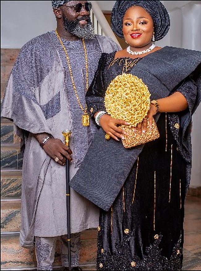 Couple in coordinated outfits: man in silver-grey embroidered agbada and cap, woman in black velvet iro and buba with navy gele