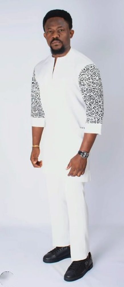 Man in a white senator with black-and-white symbol-print sleeves and white trousers on a grey studio background