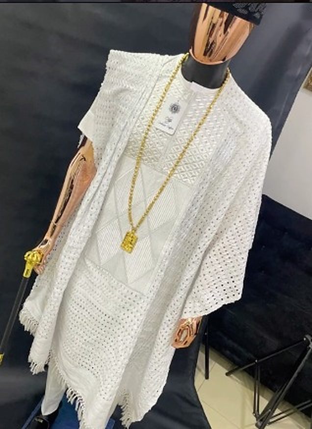 White eyelet-lace agbada with fringed hem, worn with a gold chain pendant on a mannequin