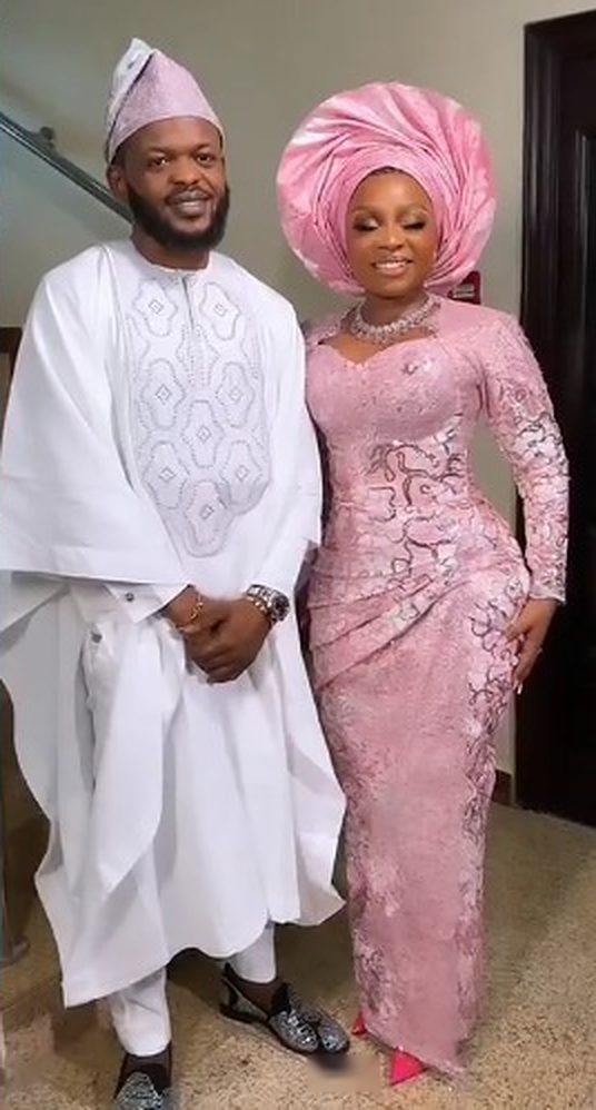 Couple in wedding attire: man in white embroidered agbada and cap, woman in blush corded-lace gown with matching gele