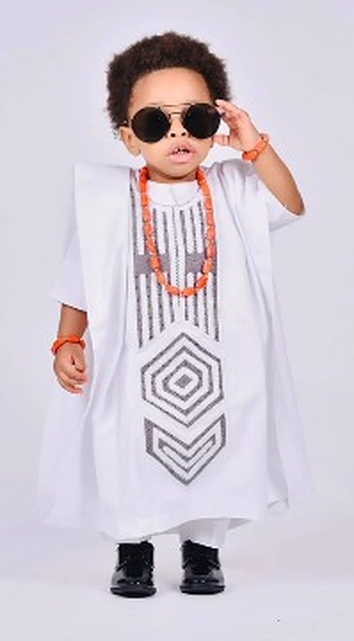 Little boy in a white agbada with silver geometric embroidery, coral bead necklace and black shoes, on a white studio background