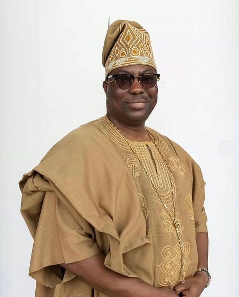 Smiling man in a camel agbada with gold-thread embroidery and matching embroidered fila, on a white studio background